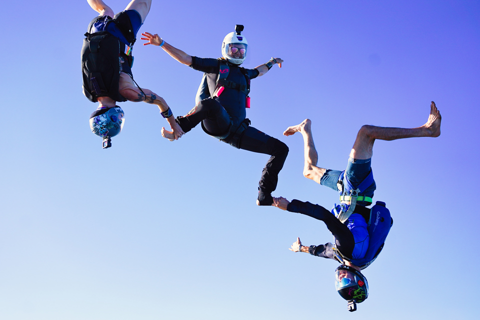 Smiling female skydiver in white helmet holds a sit position while two head down freeflyers each grip one of her feet