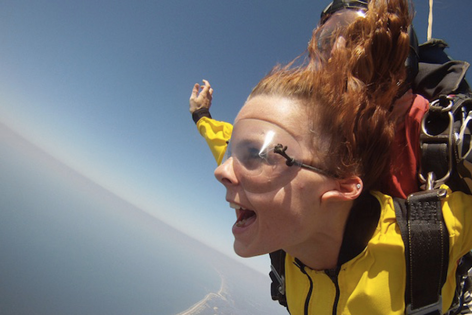 Side profile of a female tandem skydiving student in a yellow jump suit spreading her arms and enjoying freefall