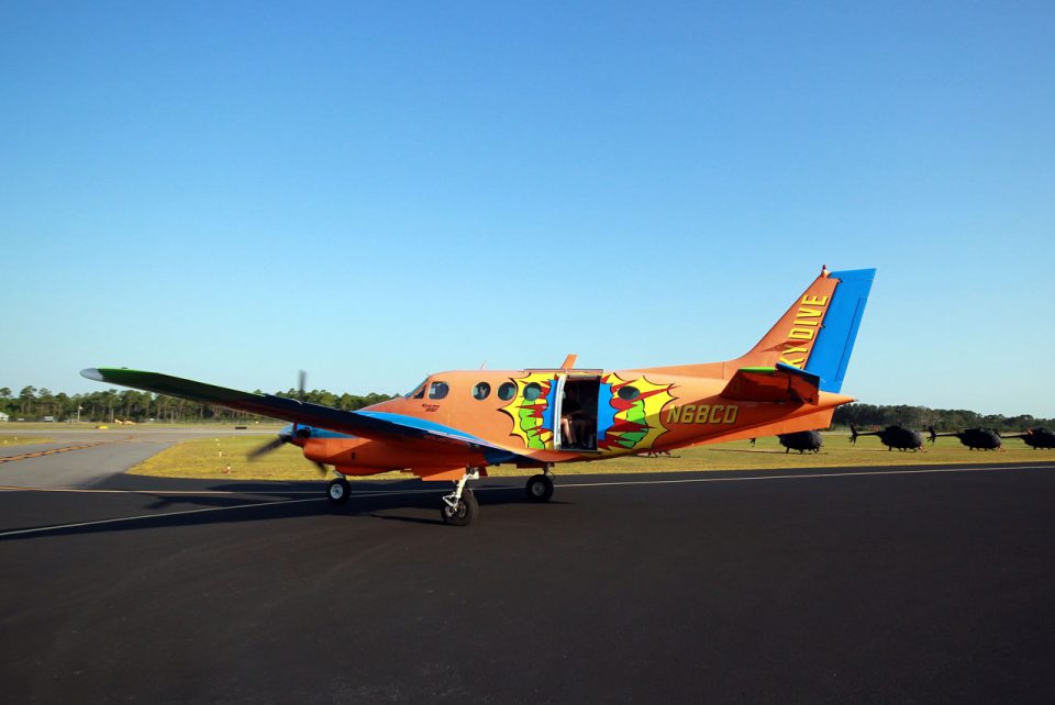 Beechcraft King Air B90 with vibrant orange paint job on runway with a row of black helicopters in the background