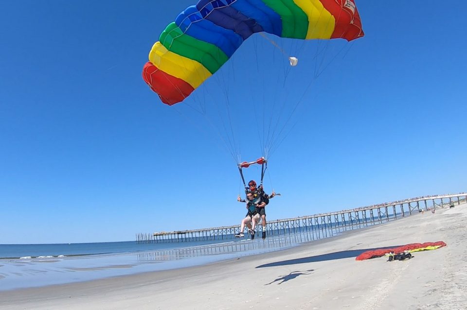 Tandem skydiving pair beneath a rainbow colored parachute landing on the beach