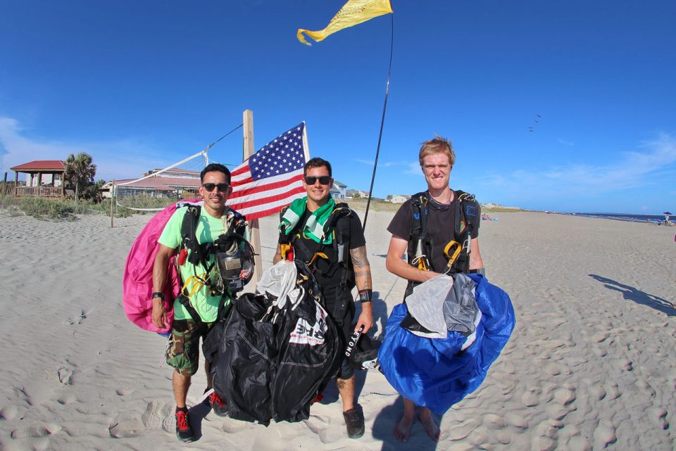 Three skydivers with parachutes in hand stand in front of American flag and volleyball net after skydiving on the beach