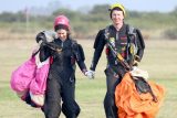 Female skydiver in pink helmet and male skydiver in yellow helmet holding hands as they walk in from the landing area