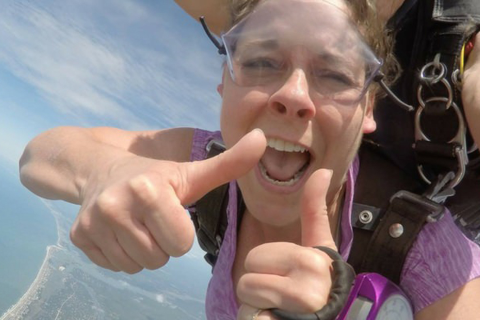 Female tandem skydiving student in clear goggles with purple analog altimeter on her hand gives thumbs up