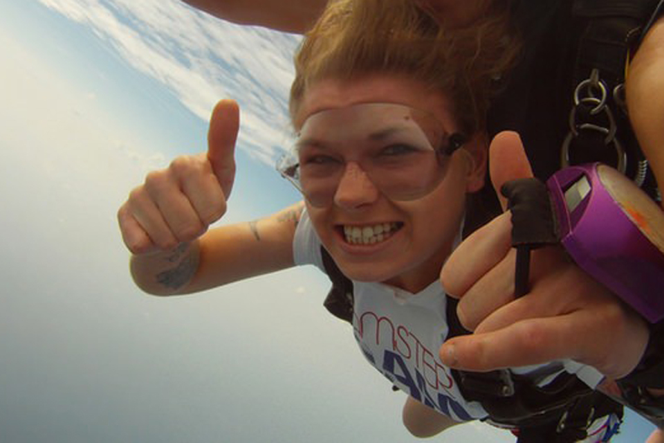 Female tandem skydiving student in clear goggles with purple analog altimeter on her hand gives thumbs up