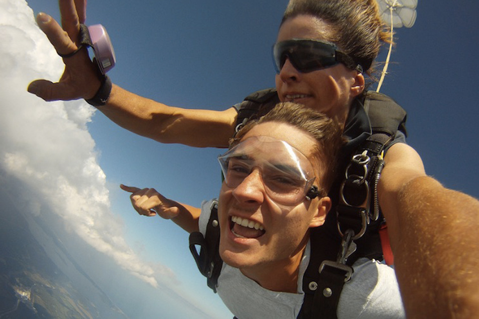 Tandem skydiving student makes the shaka gesture with one hand as female skydiving instructor in dark goggles checks altitude on an analog skydiving altimeter