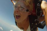 Blonde female tandem skydiving student in clear goggles letting out an excited whoop in freefall