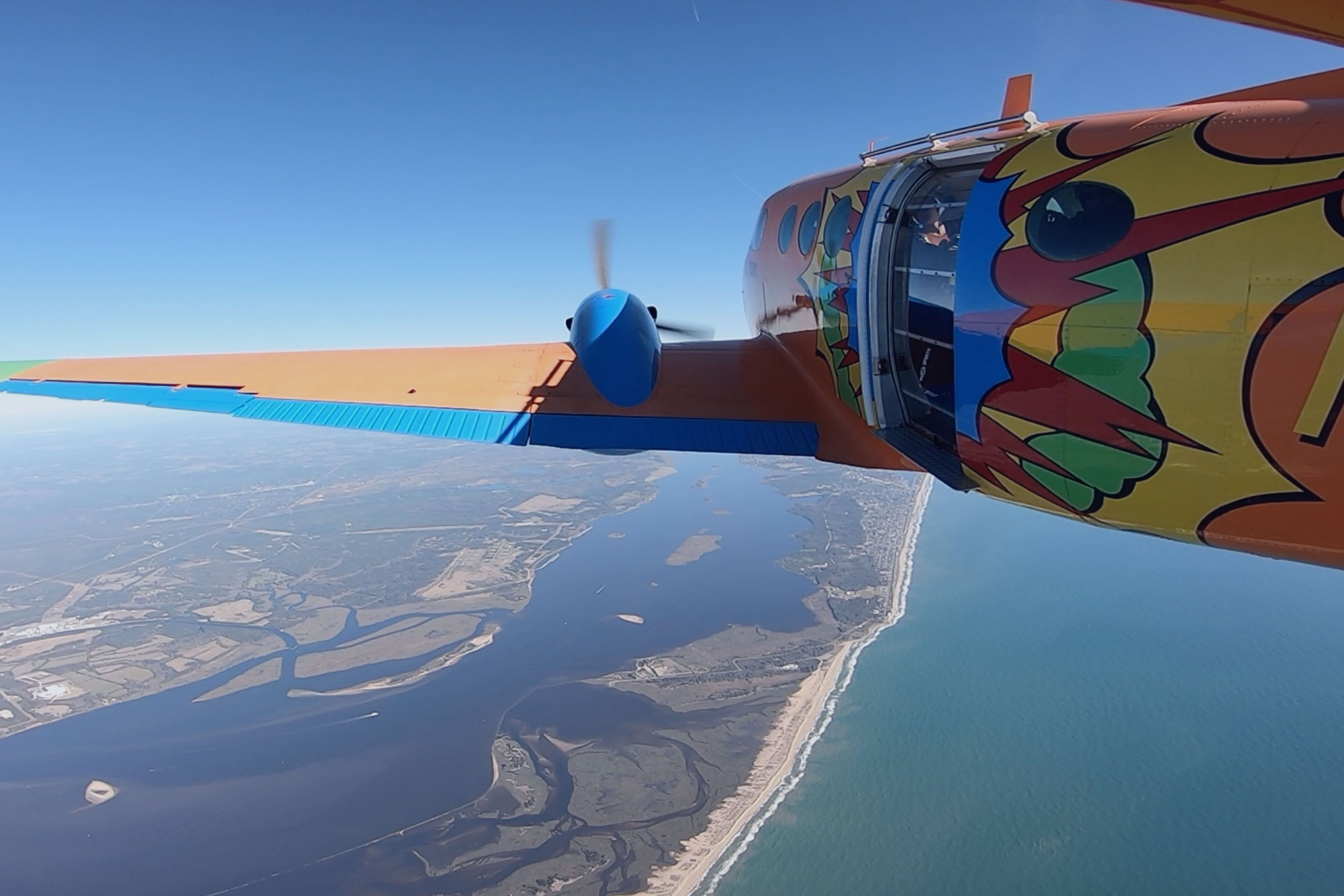 Beechcraft King Air with vibrant orange, blue, red, yellow and green pop art paint job flying over the coast of Oak Island