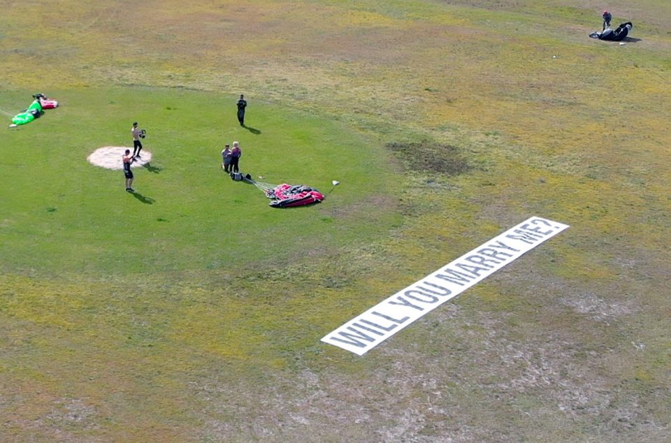 Three skydivers in the landing area record as man on bended knee proposes. There is a sign that says "will you marry me?" in the foreground. A fourth skydiver in the top right corner collects his parachute from the ground.