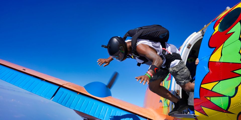 Athletic male skydiver with black helmet exiting Beechcraft King Air with bright orange, blue, red, yellow and green paint job