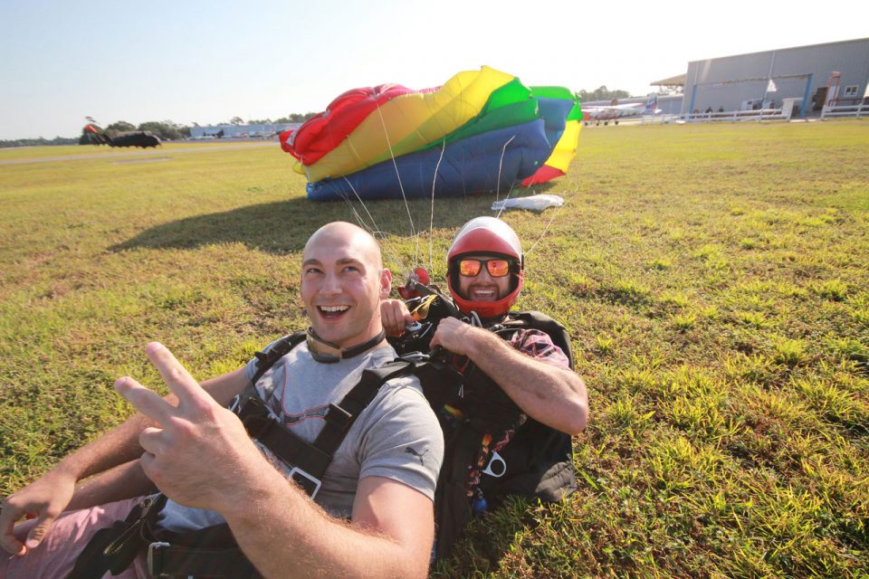 Bald tandem student giving a peace sign as smiling skydiving instructor in red helmet disconnects the harness after Myrtle Beach skydiving