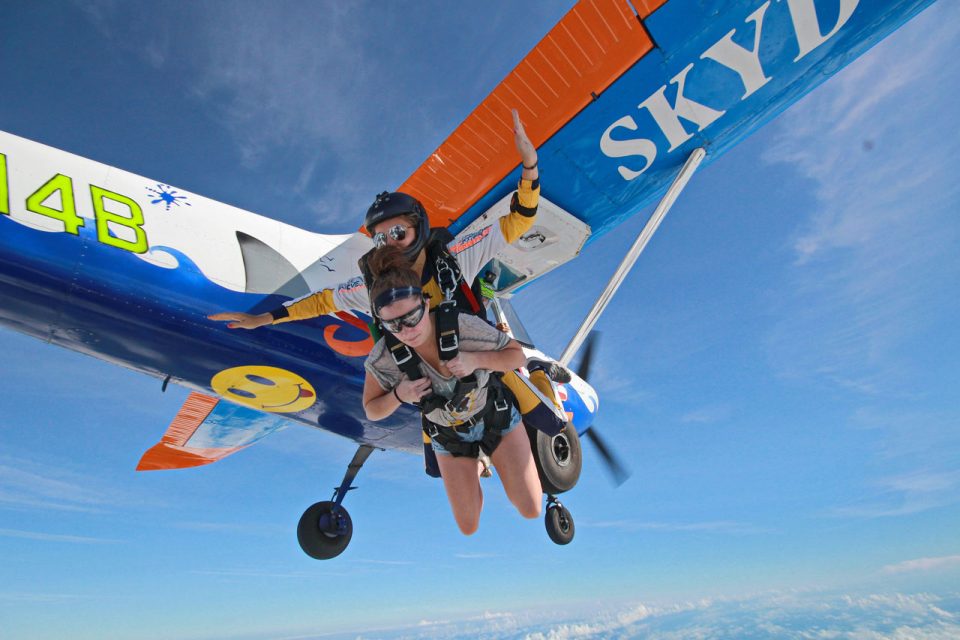 Female tandem skydiving instructor and female tandem student exit Cessna-182 painted with blue waves