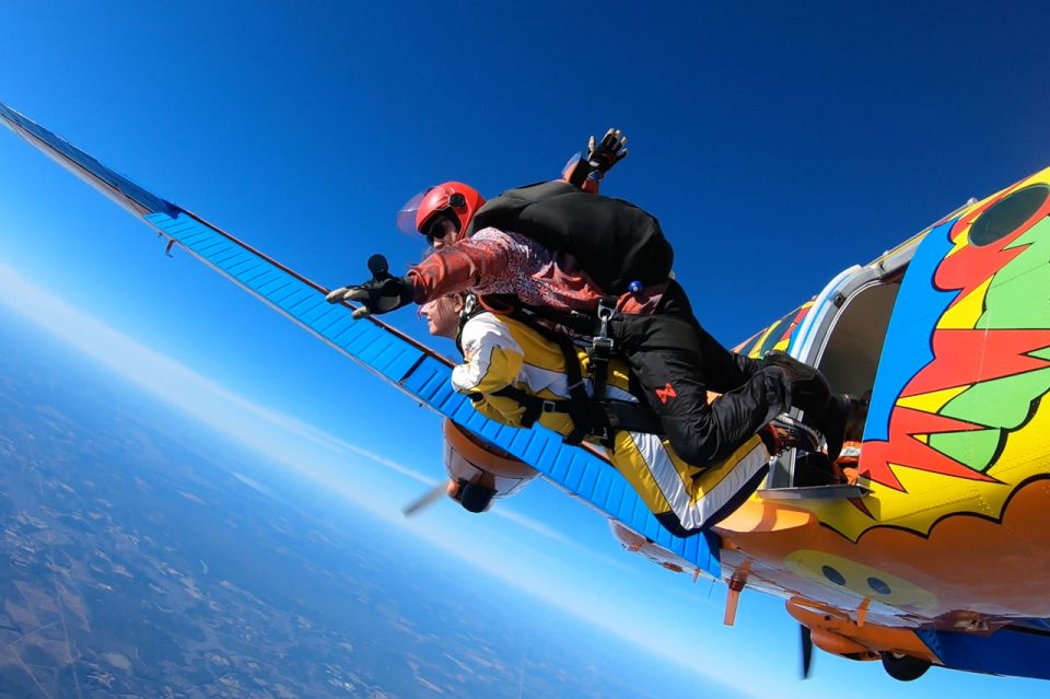 Tandem skydiving instructor wearing a red helmet with open visor using a hand cam to film the tandem student as they exit brightly painted Beechcraft King Air