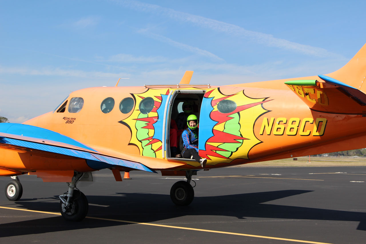 Smiling skydiver in green helmet sitting with leg hanging out of the door of the parked King Air before it takes off