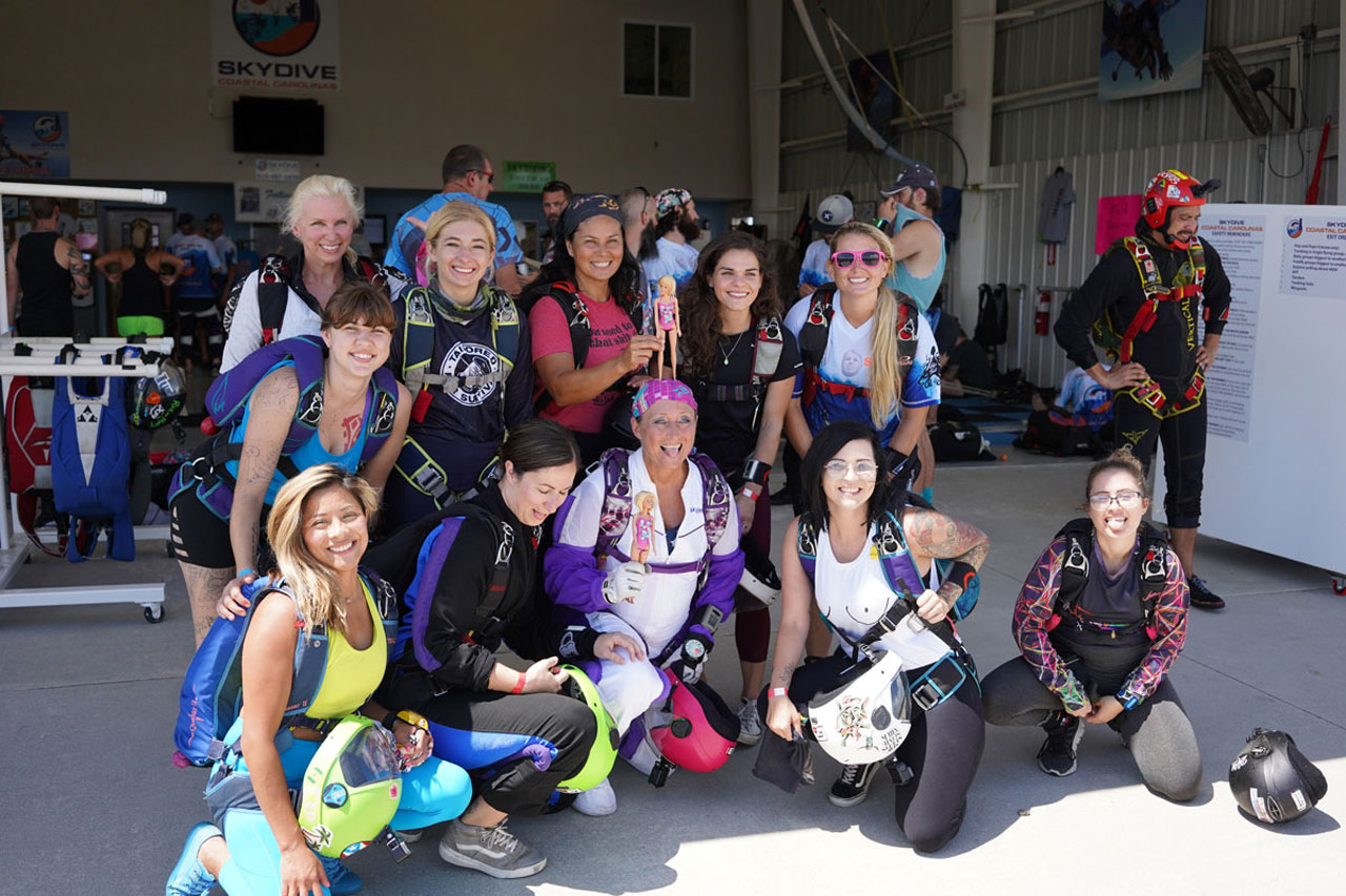 Eleven female skydivers posing with smiles and tongues out. The center two female skydivers are holding barbies.