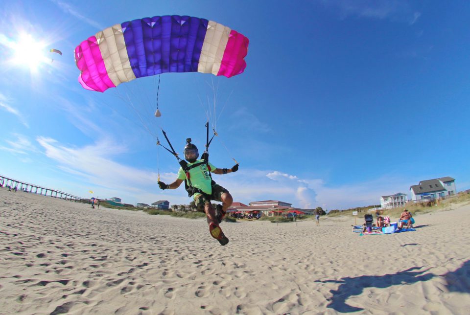 Licensed skydiver coming in for landing on the beach beneath a pink, white, and purple striped parachute