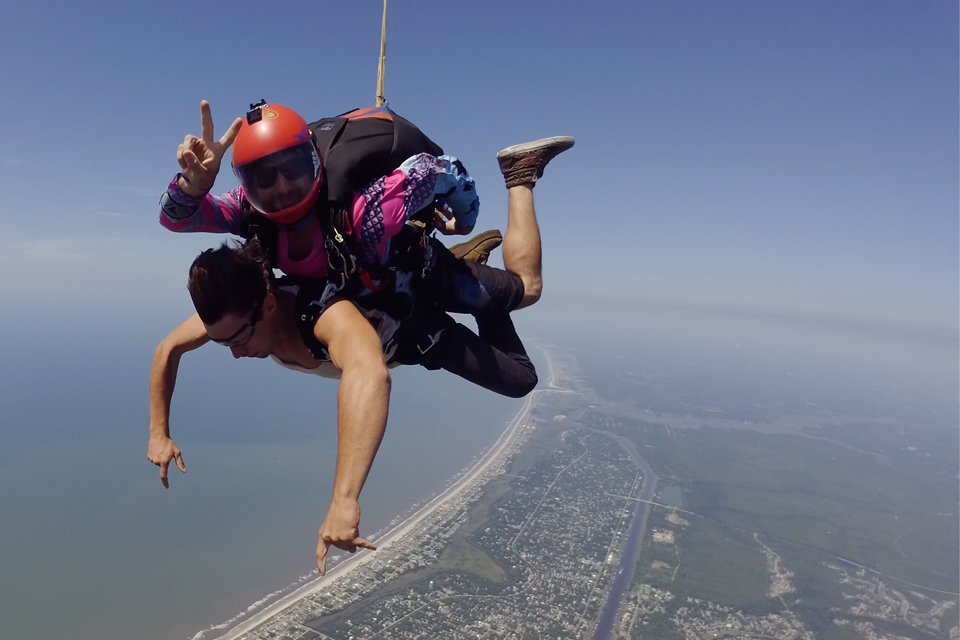 Tandem student pointing at ground while tandem skydiving instructor gives a peace sign during freefall over Oak Island, NC