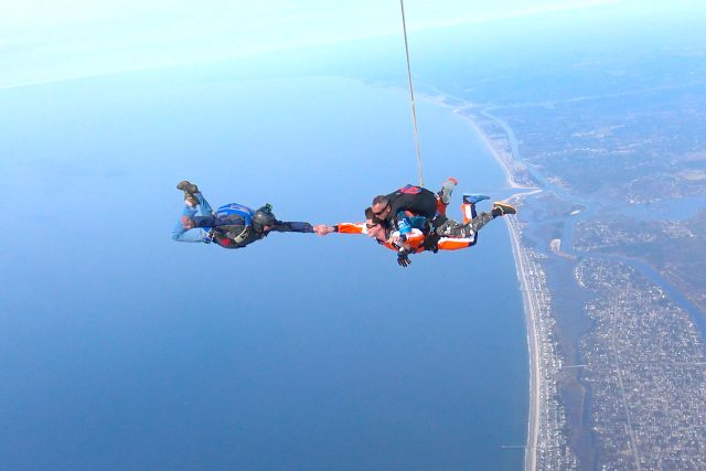 Skydiving videographer takes the hand of a tandem skydiving student over the coast of Oak Island, NC