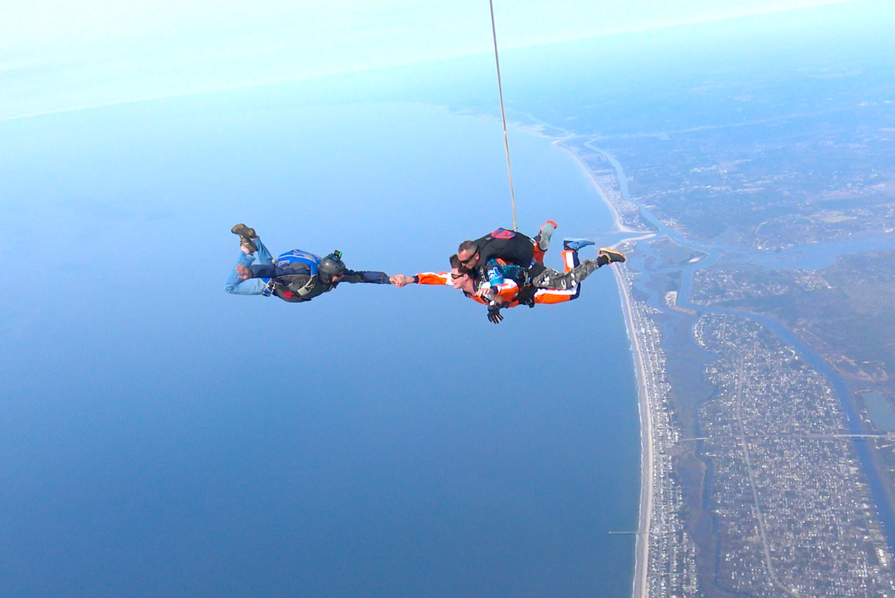 Skydiving videographer takes the hand of a tandem skydiving student over the coast of Oak Island, NC