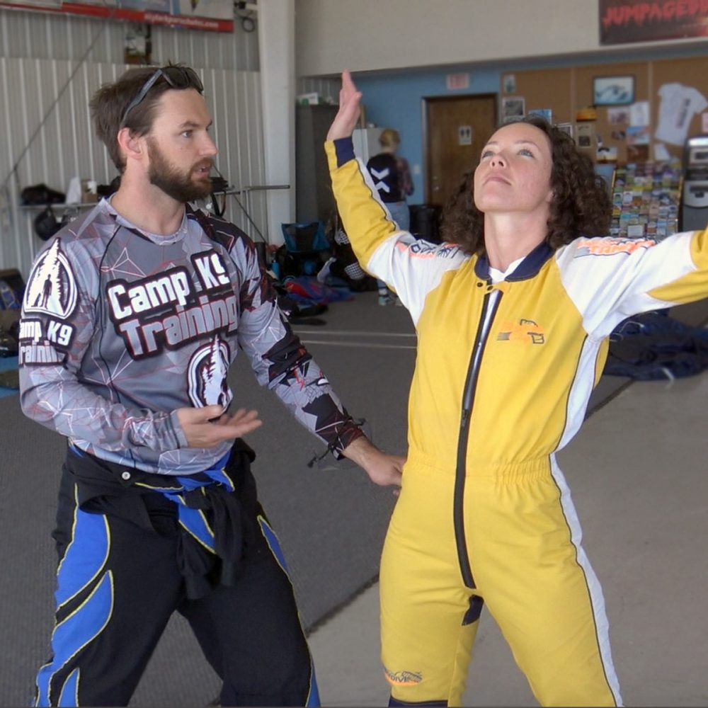 Curly haired female skydiving student in yellow jump suit practices her arch body position as skydiving instructor provides instruction