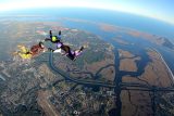Three licensed skydivers hold hands in a round formation while freefalling over the sunlit coast of Oak Island, NC