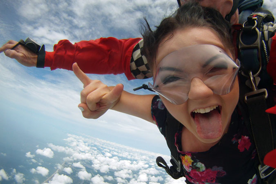 Brunette tandem skydiving student in floral top sticks out tongue and gives rock on symbol with hand while in freefall