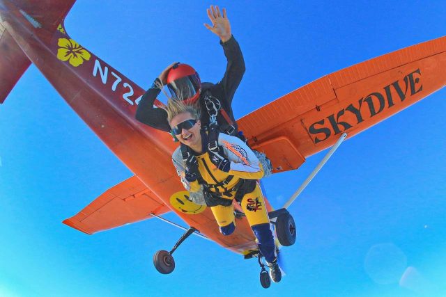 A tandem skydiving student and instructor moments after they exit the orange Cessna 182 aircraft named bubbles. The underside of the aircraft has a yellow smiley face with its tongue sticking out and the word skydive under the wing.