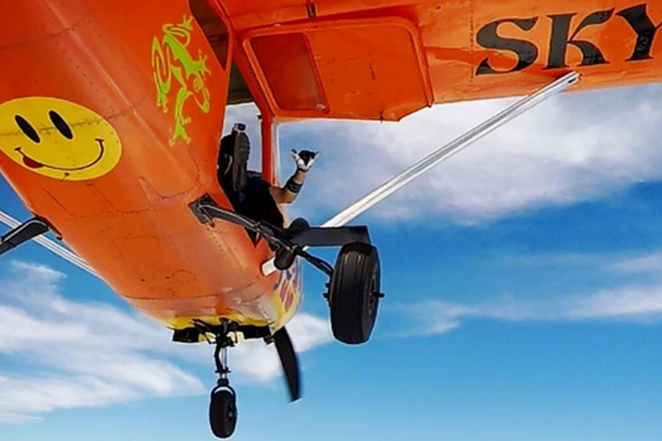 Underside view of orange Cessna-182 with skydiver seated in door giving shaka gesture with left hand
