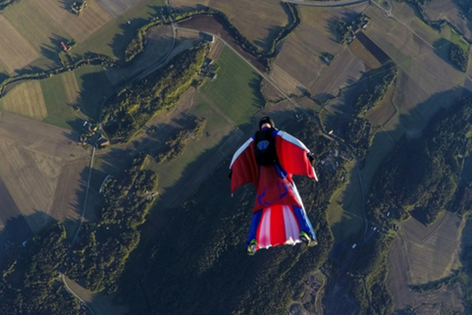 A licensed skydiver in a red white and blue wingsuit flies over a patchwork of green fields