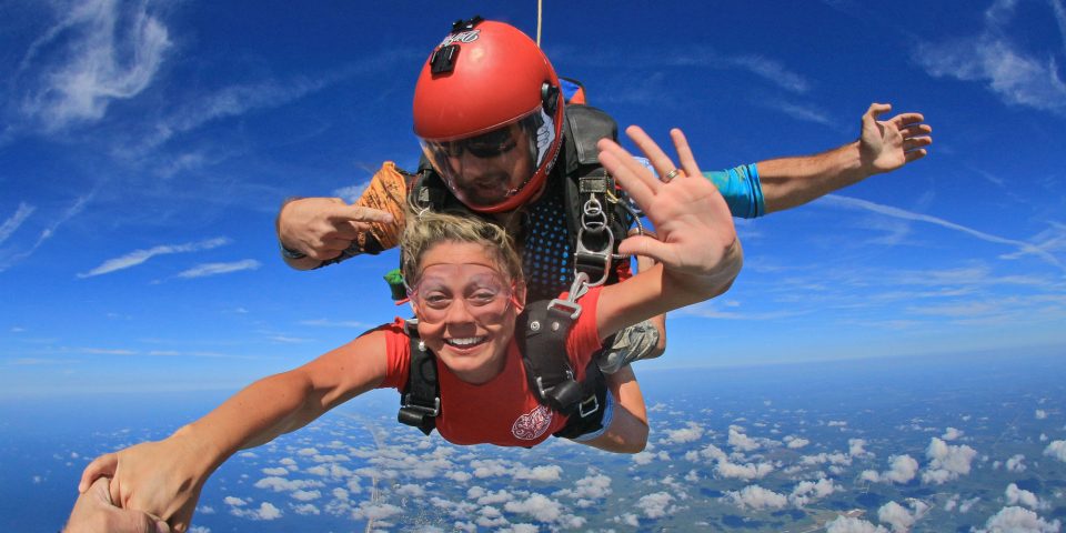 Tandem skydiving instructor in red helmet points at tandem skydiving student as she grabs the skydiving videographers hand and gestures for a high-five with the other during freefall